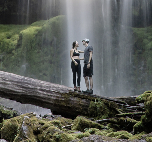 A couple standing on a fallen tree with waterfalls behind